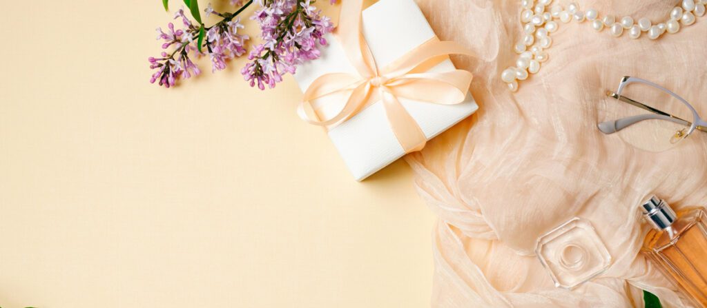 Feminine desk with silk scarf, gift box and lilac flowers on beige background. Top view, tender minimal flat lay style composition with copy space.