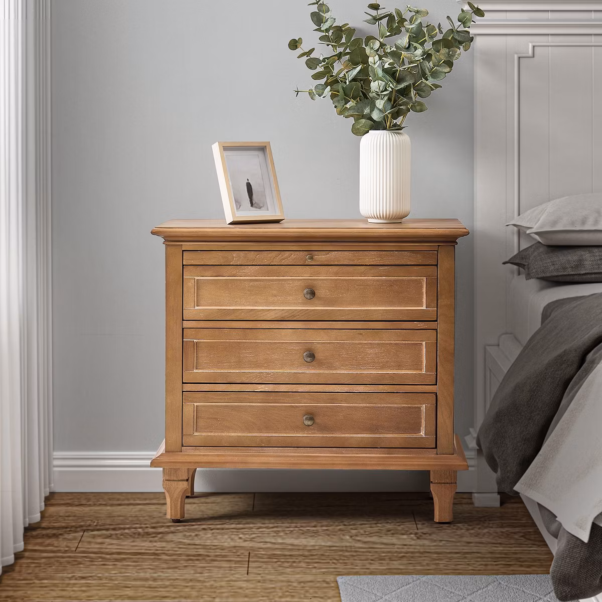 Wooden three-drawer nightstand with decor in a cozy bedroom.